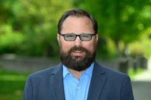 A person with a beard and glasses smiles while wearing a blue shirt and blazer, standing outdoors against a blurred green background.